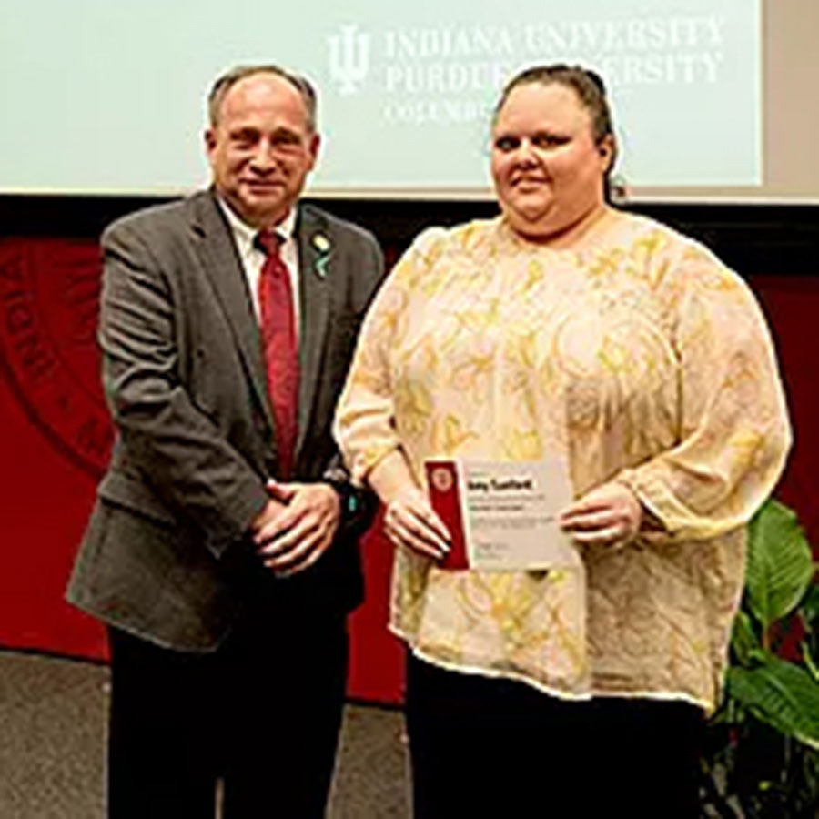 IU Columbus student smiles while holding an awards plaque between two IU educators at an Honors event.